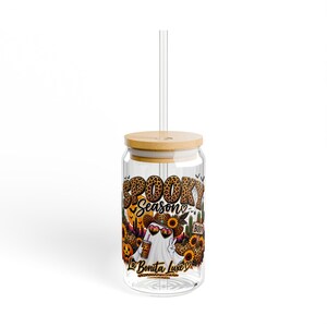 May include: A clear glass tumbler with a bamboo lid and a clear straw. The glass features a colorful graphic with the text "SPOOKY SEASON" and "La Bonita Luxe", depicting a ghost wearing a leopard print hat and sunglasses, surrounded by sunflowers, pumpkins, and cacti.