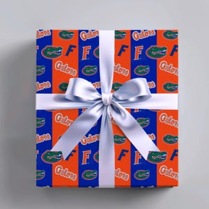 May include: A rectangular gift box wrapped in orange and blue paper featuring a repeating pattern of the Florida Gators logo, the word Gators in script, and the letter F, tied with a wide, smooth, white satin ribbon and a central bow.