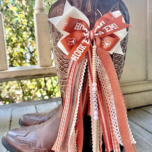 May include: A brown leather cowboy boot adorned with a decorative bow and ribbon cluster. The bow features white and burnt orange ribbons with the text HOOK 'EM and a Texas state outline. A strand of beads spells TEXAS among hanging ribbons.