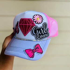 May include: A light purple trucker hat featuring a pink mesh back and a rhinestone trim. The front panel displays embroidered patches of a butterfly, a red diamond, a pink daisy, a pink bow, and the text Girl Mama in black and white script.