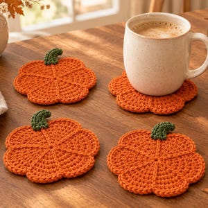 Könnte beinhalten: Ein Set aus vier orangefarbenen, kürbisförmigen Häkeluntersetzern, die jeweils mit einem kleinen grünen Stiel versehen sind. Ein Untersetzer trägt eine gesprenkelte, cremefarbene Keramiktasse, die mit Kaffee gefüllt ist.