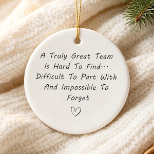 Puede incluir: Un adorno redondo de cerámica blanca con un cordón dorado para colgar. El texto dice: A Truly Great Team Is Hard To Find... Difficult To Part With And Impossible To Forget, acompañado de un pequeño símbolo de corazón dibujado a mano.