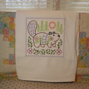 May include: White tea towel with an embroidered design of a brown bear with a purple nose and flowers in a forest setting. The bear is surrounded by green trees, a yellow sun, and purple flowers.