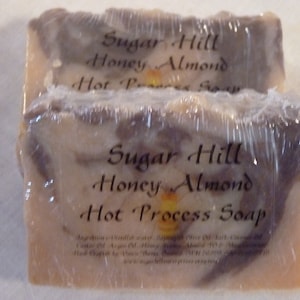 May include: Two bars of Sugar Hill Honey Almond Hot Process Soap wrapped in plastic. The soap is a light brown color with swirls of dark brown. The label is black with white text.