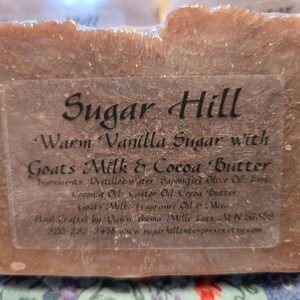 May include: A light brown rectangular soap bar, wrapped in clear plastic. The label reads "Sugar Hill" and lists ingredients including goat's milk and cocoa butter. The soap is scented with warm vanilla sugar.