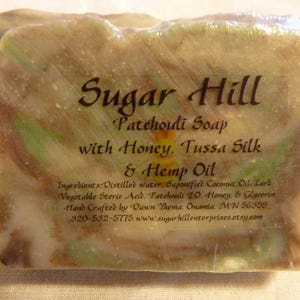 May include: A bar of Sugar Hill Patchouli Soap with Honey, Tussa Silk, and Hemp Oil. The soap is wrapped in clear plastic and features a blend of brown, green, and white colours. The label includes the soap's ingredients and maker's information.