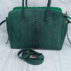 Peut inclure: Un sac à main vert foncé avec un motif texturé rappelant la peau de serpent, doté de deux anses tressées et d'une bandoulière assortie amovible avec des détails dorés. Le sac présente une forme rectangulaire structurée et une doublure intérieure verte.