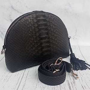 May include: A black semi-circular handbag featuring a textured snakeskin pattern, a top zipper closure, and a matching detachable shoulder strap. A black leather tassel hangs from the side, and the hardware is finished in a metallic silver tone.