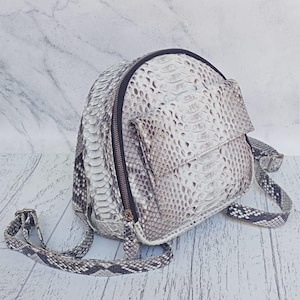 May include: A dome-shaped backpack featuring a textured snakeskin pattern in shades of white, gray, and black. The bag includes a front flap pocket, a black zipper closure with metallic hardware, and adjustable shoulder straps with a matching snakeskin print.