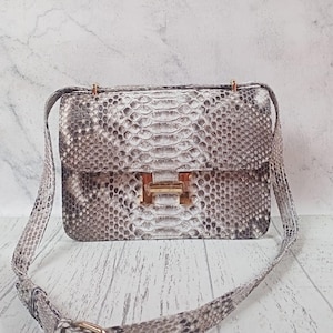 May include: A rectangular shoulder bag featuring a grey and white snakeskin pattern. The bag has a flap closure secured with a gold-toned metal H-shaped clasp. It includes an adjustable shoulder strap with a gold-toned metal buckle and hardware.