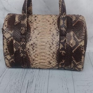Puede incluir: Un bolso de mano cilíndrico con un patrón de piel de serpiente texturizado en tonos marrón oscuro y crema. El bolso incluye dos asas a juego de piel de serpiente unidas a la parte superior, con un panel central que muestra un patrón de escamas más grande.