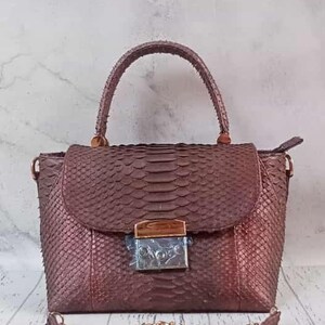 Puede incluir: Un bolso estructurado con un acabado texturizado de piel de serpiente en color bronce metálico. Cuenta con un asa superior, una solapa con un cierre metálico rectangular y una correa de hombro de cadena dorada desmontable.