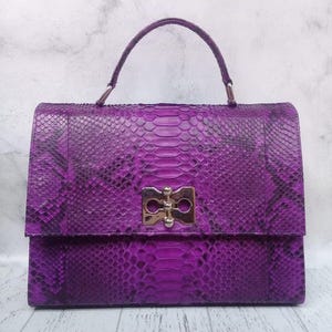 May include: A structured handbag featuring a vibrant purple snakeskin pattern with black scale accents. The bag includes a single top handle and a rectangular metallic gold-tone clasp closure centred on the front flap, showcasing a polished finish.