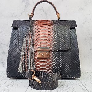 May include: A structured handbag featuring a textured snakeskin pattern in black and copper tones. The bag includes a top handle, a central metallic clasp, a decorative hanging tassel, and a detachable matching shoulder strap with gold hardware.
