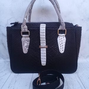 Puede incluir: Un bolso de mano negro con un patrón texturizado de piel de serpiente, que presenta dos asas con patrón de piel de serpiente gris, una correa central vertical con patrón de piel de serpiente blanca con una hebilla dorada y una correa de hombro negra extraíble con herrajes dorados.