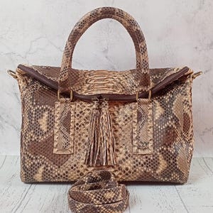 Peut inclure: Un sac à main structuré présentant un motif en peau de serpent marron et beige avec une finition texturée en écailles. Le sac comprend deux poignées supérieures, une fermeture éclair avec deux pompons assortis et une bandoulière amovible.