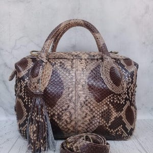 May include: A structured handbag featuring a snakeskin pattern in shades of brown, beige, and cream. The bag includes two top handles, a decorative tassel attached to one side, and a detachable, coiled shoulder strap with brass-toned hardware.