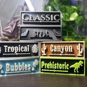 May include: A collection of five rectangular 3D-printed signs with raised lettering and icons. The signs feature the words Classic, Crypt with bats, Tropical with leaves, Canyon with cacti, Bubbles with bubbles, and Prehistoric with a dinosaur.