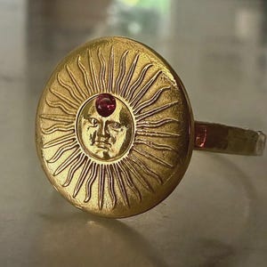 May include: A gold-toned ring featuring a circular face with an embossed sun design. The center of the sun displays a human-like face with rays extending outward. A small, round, deep red gemstone is set directly above the sun's forehead.