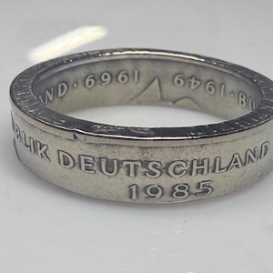 May include: A silver-toned metal ring crafted from a coin, featuring the embossed text BUNDESREPUBLIK DEUTSCHLAND and the year 1985 on the outer band. The inner band displays the text 1969 and 1949 along with a small mountain range graphic.