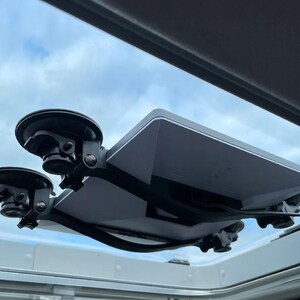 May include: A rectangular white electronic device mounted to a glass surface with four black suction cup brackets. The device features a black frame and a thin black cable extending from the side, positioned against a bright sky with light clouds.