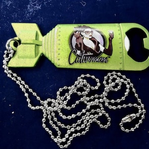 May include: A green metal bottle opener shaped like a bomb with a silver chain. The bottle opener has a graphic of Catwoman with the text "Catwoman" on it.