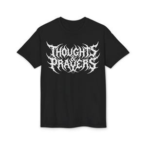 以下が含まれることがあります： 胸元に白い文字で「THOUGHTS & PRAYERS」とプリントされた黒の半袖Tシャツ。ヘヴィメタルバンドのタイポグラフィを思わせる、複雑で棘のようなディテールを持つ、鋭く尖ったスタイリッシュなフォントが特徴です。