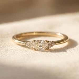 Peut inclure: Une bague en or avec une pierre centrale taille ovale, entourée de trois petites pierres rondes de chaque côté. L'anneau est fin et poli, présentant une teinte métallique chaude. Les pierres sont claires et facettées, captant la lumière.