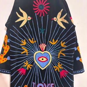 May include: A black jacket featuring vibrant, multi-colored embroidery. The design includes a central blue heart with an eye, surrounded by radiating light blue lines, yellow birds, flowers, cacti, and a moth. The word LOVE is stitched in pink.