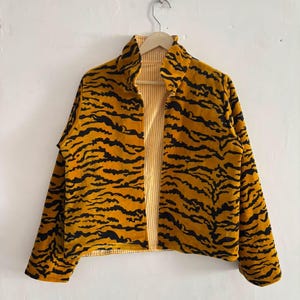 Pode incluir: Um casaco de manga comprida com um padrão de riscas de tigre em amarelo mostarda e preto no exterior. O forro interior é visível na abertura frontal e na gola, exibindo um design de riscas verticais finas em amarelo claro e branco.