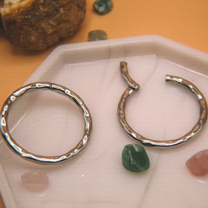 May include: Two silver-toned circular hoop rings with a textured, hammered surface. One ring is closed, while the other is open, revealing a hinged segment mechanism designed for secure closure. The metal has a reflective, polished finish.