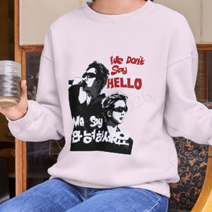 Puede incluir: Una sudadera de manga larga de color rosa claro con un estampado gráfico de dos personas con gafas de sol. El texto dice "We Don't Say HELLO We Say 안녕하세요" en tinta roja y negra. La prenda tiene cuello redondo y puños acanalados.