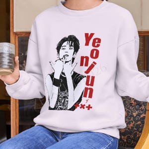 May include: A white long-sleeve sweatshirt featuring a black and white graphic illustration of a person with dark hair making a hand gesture. The text "Yeonjun" is printed in bold red vertical lettering next to the graphic, with "+x+" below it.