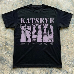 May include: A black short-sleeve t-shirt featuring the word KATSEYE in large light purple letters at the top. Below the text are six individual black and white portrait panels of people, each with a handwritten-style signature underneath them.