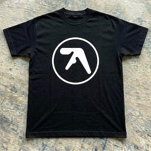 May include: A black short-sleeved t-shirt featuring a large white graphic print on the chest. The graphic consists of a stylized, abstract geometric logo enclosed within a solid white circle. The shirt has a crew neckline and a standard fit.