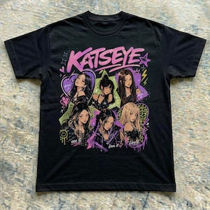 May include: A black short-sleeve t-shirt featuring a graphic print of the group KATSEYE. The design includes six illustrated figures in purple and pink tones, surrounded by stars, hearts, and text that reads dream it, touch it, own it, see it, love it, live it.