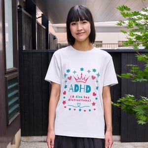 May include: A white short-sleeved t-shirt featuring a graphic design with a pink crown, teal text reading ADHD, and the phrase (A diva has distractions) in pink. The design is surrounded by small teal and pink hearts, stars, and flower icons.