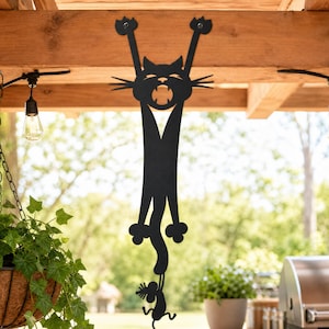 May include: A black metal wall art piece featuring a stylized cat hanging by its front paws with its mouth open, while its long tail curls downward to hold a small mouse figure. The silhouette is mounted vertically on a wooden beam structure.