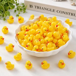 Op de afbeelding: Een grote witte kom met een geschulpte rand gevuld met veel kleine gele rubberen eendjes, met extra individuele gele eendjes verspreid rond de basis van de kom. Elk eendje heeft een felgeel lichaam en een kleine oranje snavel.