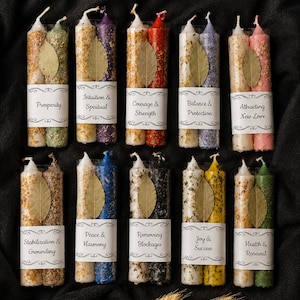 May include: Ten pairs of cylindrical candles, each wrapped in a white paper band with printed text and a dried leaf. The candles feature various colors including gold, green, purple, red, blue, black, yellow, and pink, with textured surfaces.