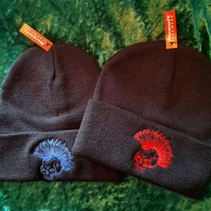 Peut inclure: Deux bonnets en tricot noir avec un motif de crâne et de mohawk. Un bonnet a un mohawk bleu et l'autre a un mohawk rouge.