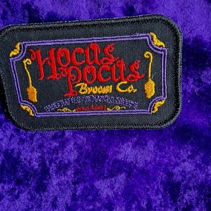 Peut inclure: Patch brodé noir avec le texte "Hocus Pocus Broom Co." en lettres rouges et dorées. Le patch présente deux balais dorés.
