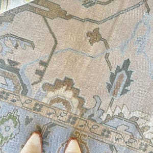 May include: A woven area rug featuring a light beige background with a repeating pattern of floral and geometric motifs in shades of dusty blue, sage green, and tan. The rug has a visible border and a pair of light-colored leather boots.