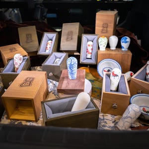 May include: A collection of ceramic stamps with various patterns including blue and white floral designs, red floral motifs, and rooster illustrations, each housed in individual wooden boxes with grey fabric linings and branded logos.