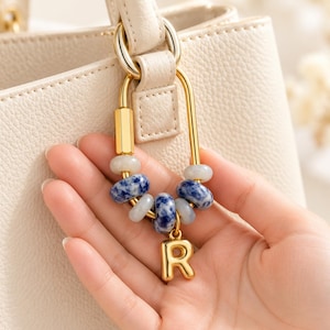 May include: A gold-toned metal carabiner bag charm featuring a series of round beads in shades of blue, white, and grey, accented with a polished gold-toned letter R charm. The accessory is attached to the handle of a textured cream-colored handbag.