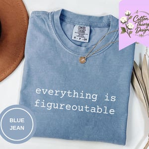May include: A blue jean colored t-shirt with the white text everything is figureoutable printed on the front. The shirt features a crew neckline and a Comfort Colors brand label on the inside collar. A gold necklace with a small round pendant rests on the fabric.