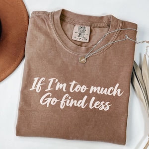 May include: A brown short-sleeve t-shirt with a crew neckline featuring the white printed text If I'm too much Go find less. The shirt has a Comfort Colors brand label on the inside collar indicating size S-CH and 100% cotton material composition.