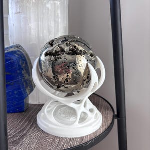 May include: A metallic pyrite sphere with a textured, crystalline surface featuring dark crevices, resting in a white, three-pronged, organic-shaped stand. The sphere has a reflective, polished finish that shows a distorted reflection of the surroundings.