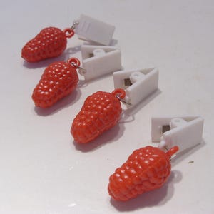Op de afbeelding: Vier rode, besvormige bedels met witte, driehoekige clips. De bedels hebben een textuuroppervlak en zijn met kleine zilveren ringen aan de clips bevestigd. De items zijn diagonaal op een wit oppervlak geplaatst.