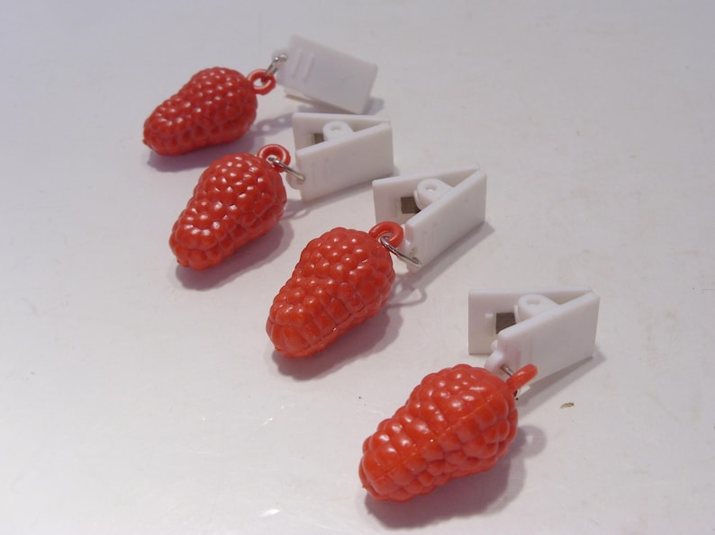 Op de afbeelding: Vier rode, besvormige bedels met witte, driehoekige clips. De bedels hebben een textuuroppervlak en zijn met kleine zilveren ringen aan de clips bevestigd. De items zijn diagonaal op een wit oppervlak geplaatst.